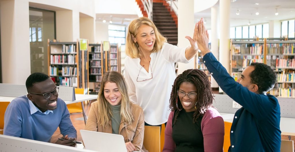five adults smile around a library table with a laptop and two people high-five