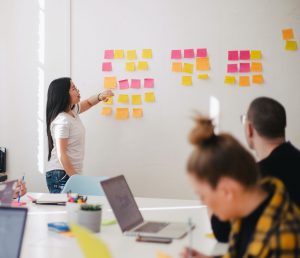 a women gesturing towards a wall of post its while other people sit at a table
