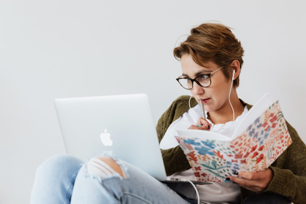 a young woman wearing glasses gazes at a laptop in thought while holding a notebook